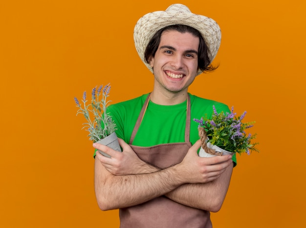 Giovane giardiniere uomo in grembiule e cappello che tiene piante in vaso guardando sorridente allegramente con le mani incrociate in piedi sopra la parete arancione