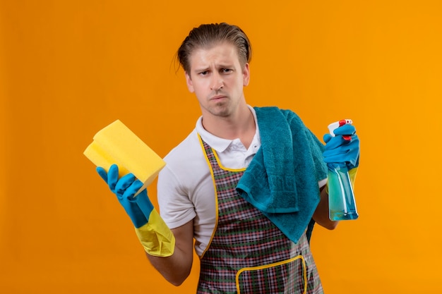 Giovane hansdome uomo che indossa un grembiule e guanti di gomma tenendo spray per la pulizia e spugna scontento con la faccia accigliata in piedi sopra la parete arancione