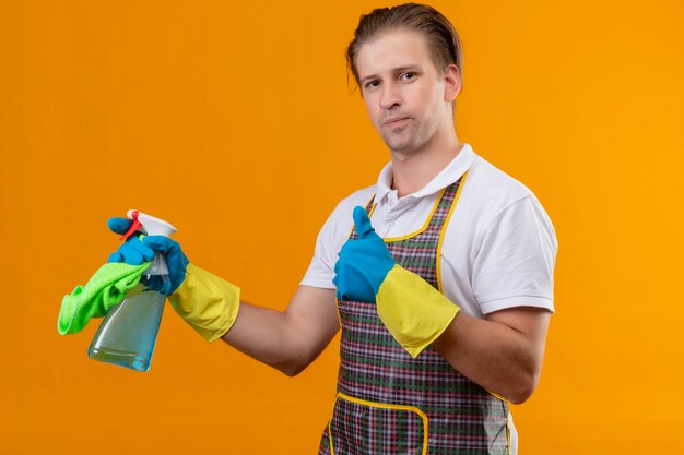 Giovane hansdome uomo che indossa un grembiule e guanti di gomma tenendo spray per la pulizia e tappeto con sorriso fiducioso sul viso che mostra i pollici in su in piedi sopra la parete arancione