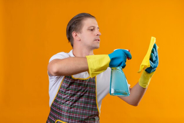 Giovane hansdome uomo che indossa un grembiule e guanti di gomma tenendo tappeto utilizzando spray detergente