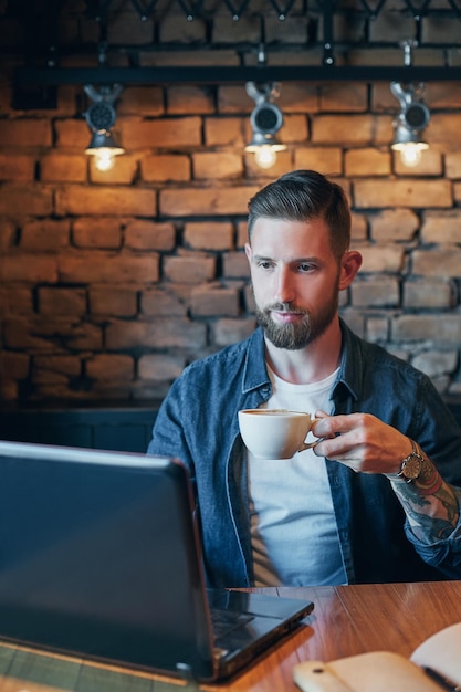 Giovane hipster al bar che beve un cappuccino, giovane che beve caffè in un caffè della città durante l'ora di pranzo e lavora sul portatile