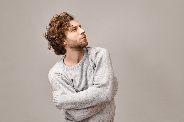 Giovane hipster alla moda alla moda con barba curata, lineamenti del viso ondulati rossastri e ideali che guardano in alto con espressione pensierosa indossando un pullover elegante oversize, tenendo le braccia incrociate sul petto