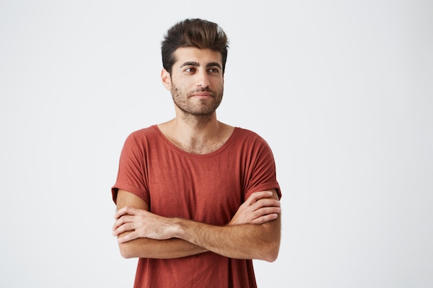 Giovane hipster spagnolo che indossa una maglietta rossa leggermente sorridente, tenendo le mani incrociate e lo sguardo meditativo da parte durante il servizio fotografico della rivista. Concetto di bellezza, persone, gioventù e stile di vita