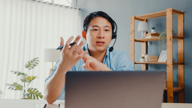 Giovane imprenditore asiatico indossare le cuffie utilizzando laptop parlare con i colleghi del piano in videochiamata mentre si lavora da casa in salotto.