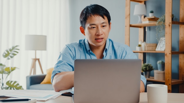 Giovane imprenditore asiatico utilizzando laptop parlare ai colleghi del piano in videochiamata mentre si lavora in modo intelligente da casa