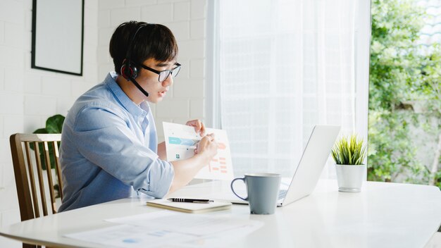 Giovane imprenditore asiatico utilizzando laptop parlare con i colleghi del piano in videochiamata mentre si lavora in modo intelligente da casa in soggiorno. Autoisolamento, allontanamento sociale, quarantena per la prevenzione del coronavirus.