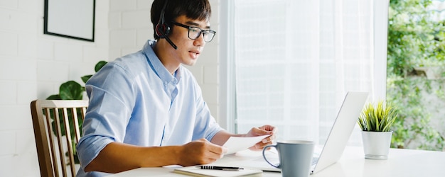 Giovane imprenditore asiatico utilizzando laptop parlare con i colleghi del piano in videochiamata mentre si lavora in modo intelligente da casa in soggiorno. Autoisolamento, allontanamento sociale, quarantena per la prevenzione del coronavirus.