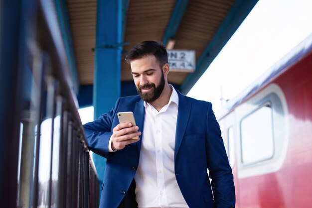 Giovane imprenditore barbuto in abito elegante in attesa del treno della metropolitana per andare al lavoro e utilizzando il suo smartphone