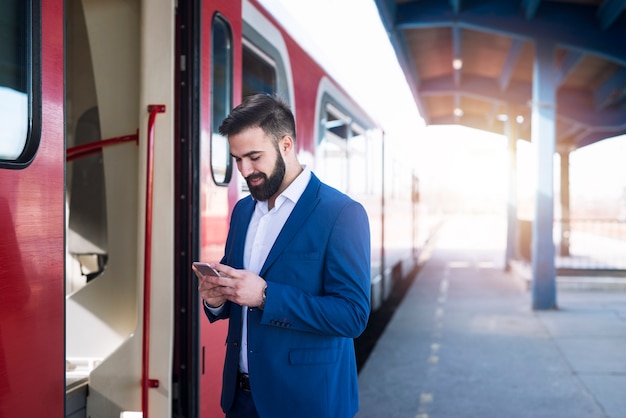 Giovane imprenditore barbuto in abito elegante in attesa del treno della metropolitana per andare al lavoro e utilizzando il suo smartphone