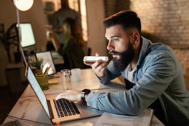 Giovane imprenditore che legge un'e-mail sul computer mentre registra un messaggio vocale sullo smartphone durante il lavoro notturno in ufficio