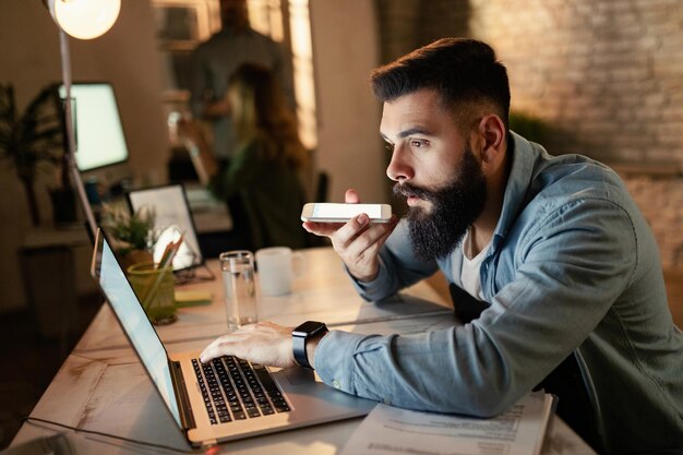 Giovane imprenditore che legge un'e-mail sul computer mentre registra un messaggio vocale sullo smartphone durante il lavoro notturno in ufficio