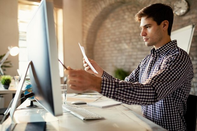 Giovane imprenditore che utilizza il computer mentre confronta i dati con i rapporti aziendali in ufficio
