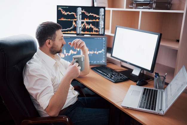 Giovane imprenditore è seduto in ufficio al tavolo, lavorando sul computer con molti monitor, diagrammi sul monitor. L'agente di borsa analizza i grafici delle opzioni binarie Uomo dei pantaloni a vita bassa che beve caffè, studiando