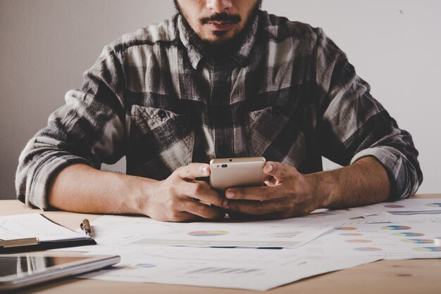 Giovane imprenditore seduto e utilizzando il telefono cellulare sul posto di lavoro in ufficio. L&#39;uomo d&#39;affari prende una pausa mentre lavora l&#39;analisi con i dati di business.