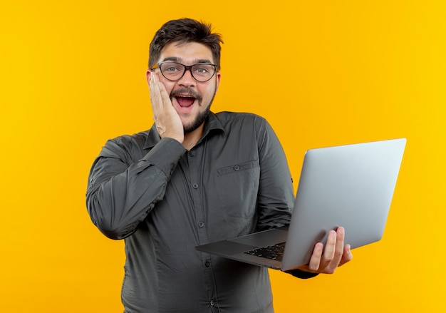 Giovane imprenditore sorpreso con gli occhiali in possesso di laptop e mettendo la mano sulla guancia isolato su sfondo giallo