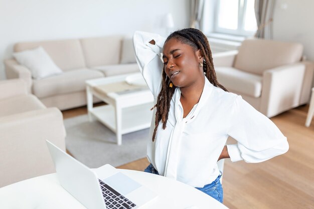 Giovane imprenditrice africana avendo mal di schiena mentre era seduto alla scrivania in ufficio Imprenditrice tenendo la schiena mentre si lavora al computer portatile alla scrivania in ufficio