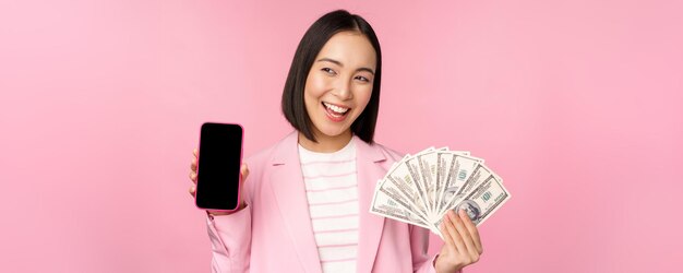 Giovane imprenditrice asiatica di successo che mostra soldi contanti dollari e schermo dello smartphone sorridente soddisfatto sfondo rosa