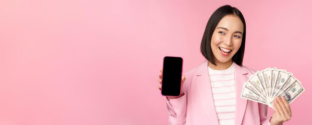 Giovane imprenditrice asiatica di successo che mostra soldi contanti dollari e schermo dello smartphone sorridente soddisfatto sfondo rosa