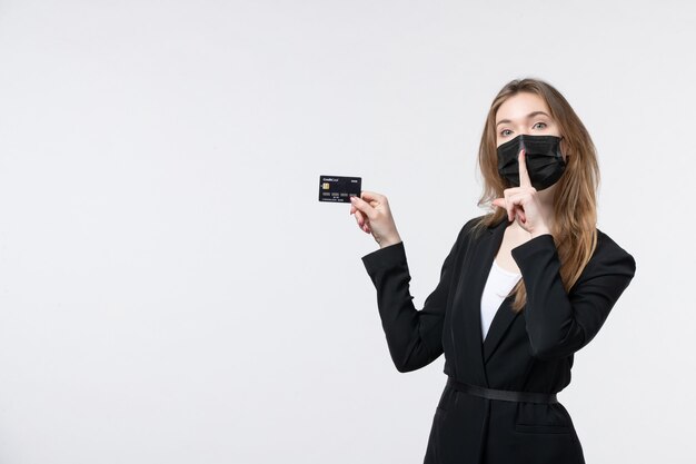 Giovane imprenditrice seria in tuta che indossa la sua maschera medica e mostra la carta di credito facendo gesto di silenzio sul muro bianco
