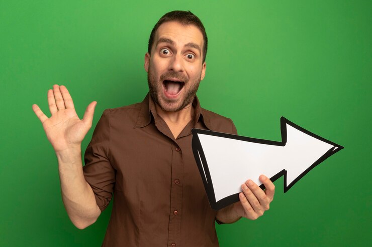 Foto gratuito giovane impressionato che guarda il segno della freccia della tenuta anteriore che indica al lato che mostra la mano vuota isolata sulla parete verde