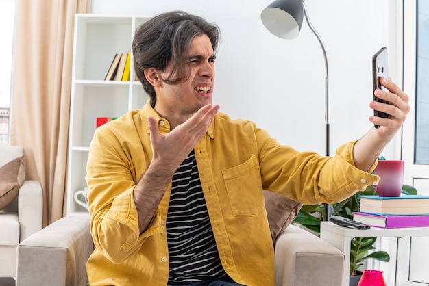 Giovane in abiti casual che fa selfie usando lo smartphone che sembra arrabbiato e frustrato seduto sulla sedia in un soggiorno luminoso