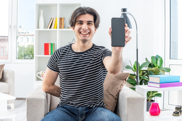 Giovane in abiti casual che mostra smartphone felice e allegro sorridente ampiamente seduto sulla sedia in un soggiorno luminoso