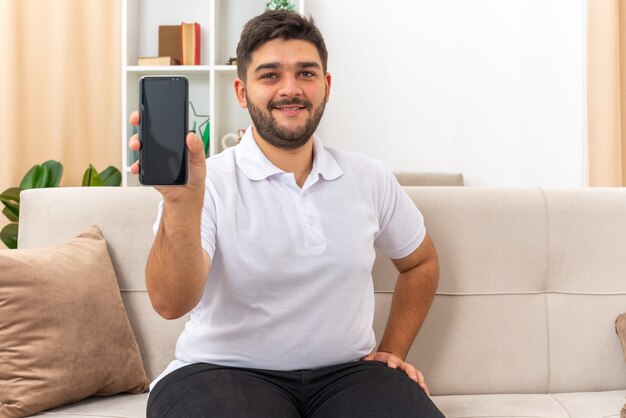 Giovane in abiti casual che mostra uno smartphone che sembra felice e positivo sorridente seduto su un divano in un soggiorno luminoso