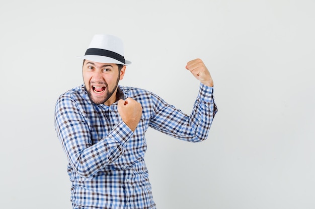 Giovane in camicia a quadri, cappello che mostra il gesto del vincitore e che sembra felice, vista frontale.