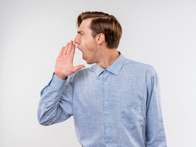 Giovane in camicia blu che osserva da parte gridando con la mano vicino alla bocca che sta sopra il muro bianco