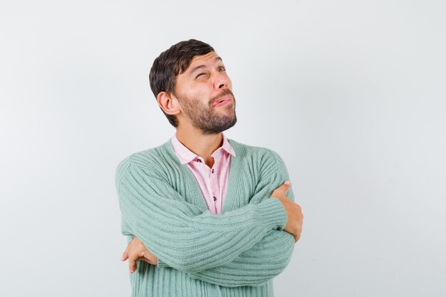 Giovane in camicia, cardigan in piedi con le braccia incrociate, alzando lo sguardo e guardando pensieroso, vista frontale.