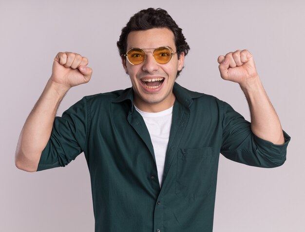 Giovane in camicia verde con gli occhiali felici ed eccitati stringendo i pugni che rallegrano il suo successo in piedi sopra il muro bianco