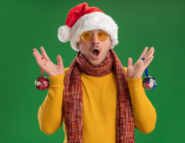Giovane in dolcevita giallo e cappello da Babbo Natale con gli occhiali che tengono i giocattoli per l'albero di Natale stupito e sorpreso in piedi sopra la parete verde