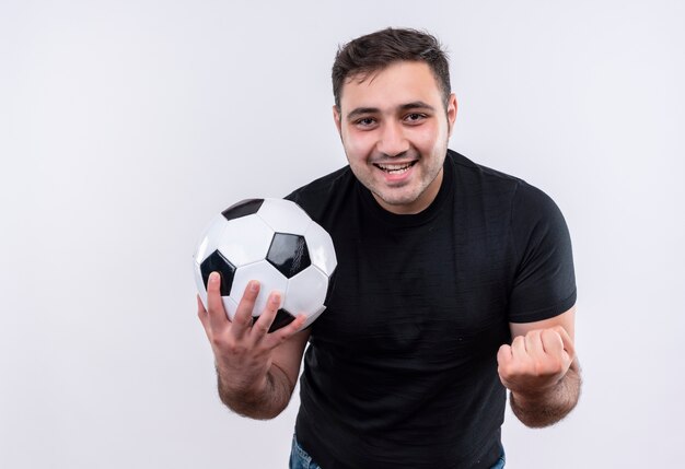 Giovane in maglietta nera che tiene calcio bal l pugno di serraggio felice ed eccitato in piedi sopra il muro bianco