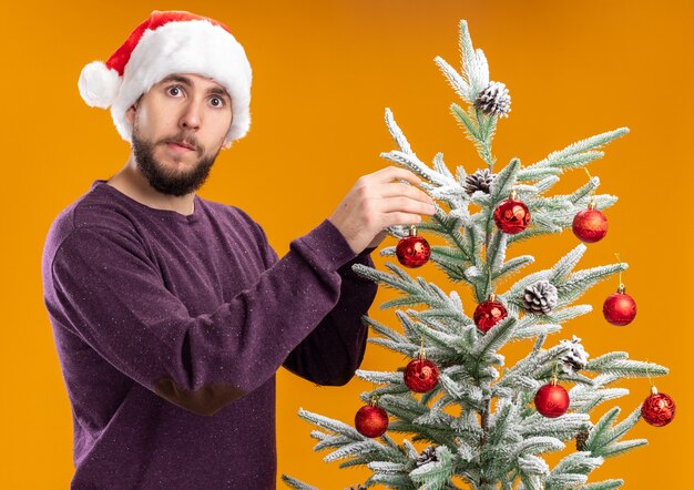 Giovane in maglione viola e cappello da Babbo Natale che appende i giocattoli sull'albero di Natale che sembra confuso in piedi sopra la parete arancione