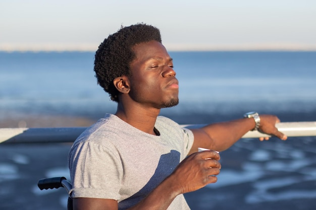 Giovane in sedia a rotelle con caffè da asporto all'aperto. Ragazzo nero che beve caffè sullo sfondo del paesaggio marino e si aggrappa alla rotaia con una mano. Stile di vita, disabilità, concetto di motivazione.