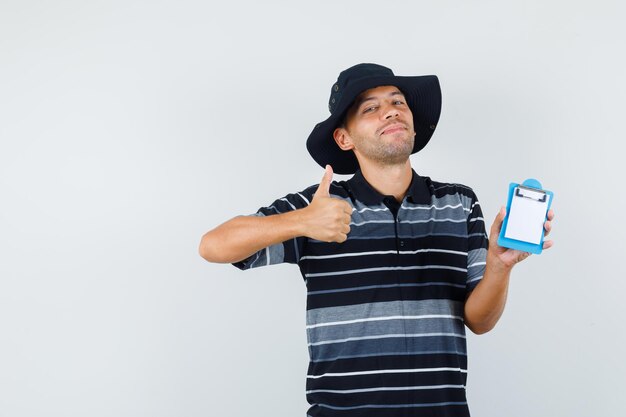 Giovane in t-shirt, cappello che tiene mini appunti con il pollice in su e sembra allegro, vista frontale.