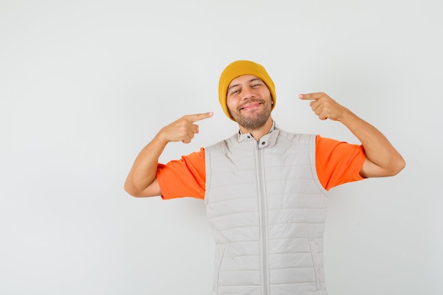 Giovane in t-shirt, giacca, cappello che punta al suo sorriso e sembra allegro