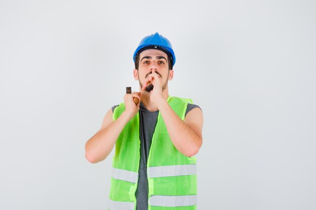 Giovane in uniforme da costruzione che alza l'ascia sopra la spalla e mostra il gesto del silenzio e sembra concentrato