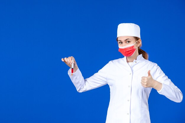 Giovane infermiera di vista frontale in vestito medico bianco con maschera rossa e iniezione nelle sue mani sulla parete blu