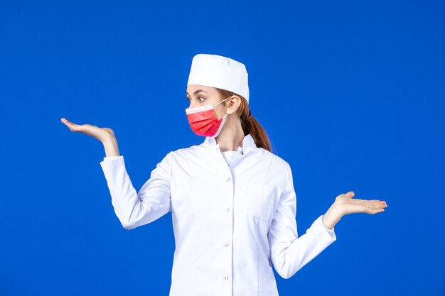 Giovane infermiera di vista frontale in vestito medico con maschera protettiva rossa sulla parete blu