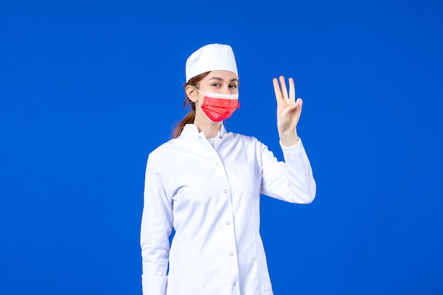 Giovane infermiera di vista frontale in vestito medico con maschera protettiva rossa sulla parete blu