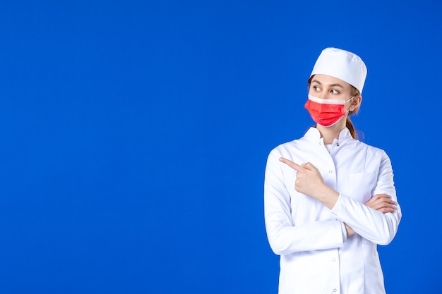 Giovane infermiera di vista frontale in vestito medico con maschera protettiva rossa sulla parete blu