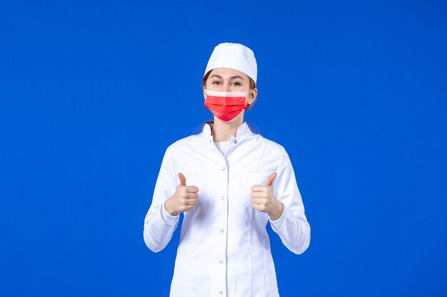 Giovane infermiera di vista frontale in vestito medico con maschera protettiva rossa sulla parete blu