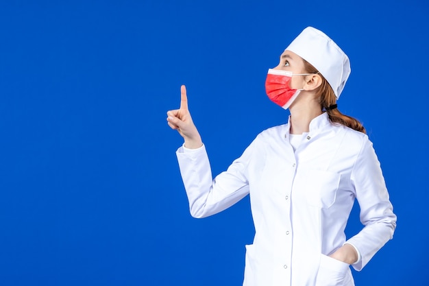 Giovane infermiera di vista frontale in vestito medico con maschera protettiva rossa sulla parete blu
