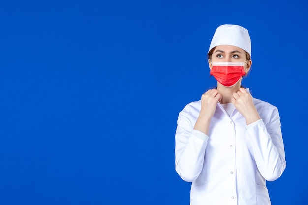 Giovane infermiera di vista frontale in vestito medico con maschera rossa sulla parete blu