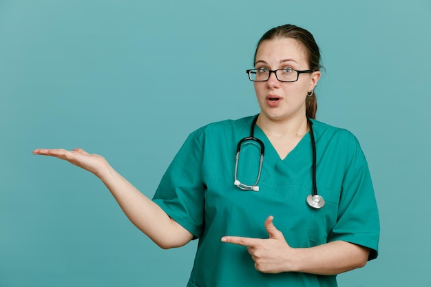 Giovane infermiera in uniforme medica con stetoscopio intorno al collo che presenta qualcosa con la mano sulla mano che punta con il dito indice di lato guardando confuso in piedi su sfondo blu