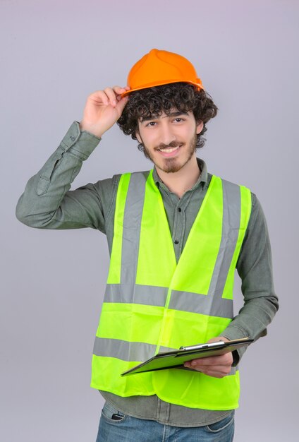 Giovane ingegnere bello barbuto che fa gesto di saluto che tocca il suo casco che sorride controllando muro bianco isolato