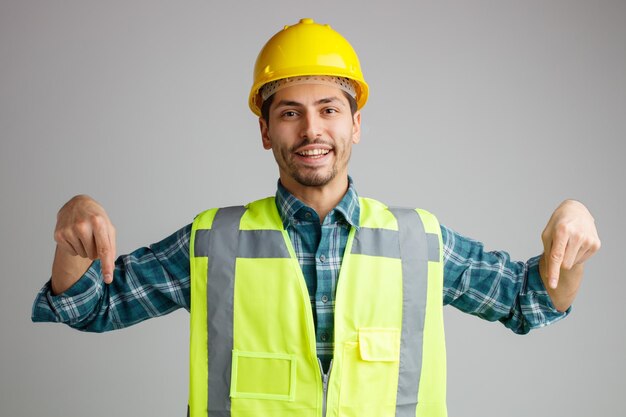 Giovane ingegnere maschio gioioso che indossa casco di sicurezza e uniforme guardando la fotocamera che punta le dita verso il basso isolato su sfondo bianco