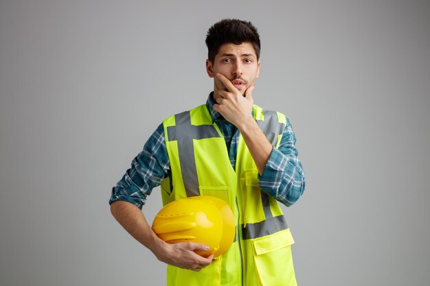 Giovane ingegnere maschio premuroso che indossa l'uniforme che tiene il casco di sicurezza guardando la fotocamera tenendo la mano sul mento isolato su sfondo bianco