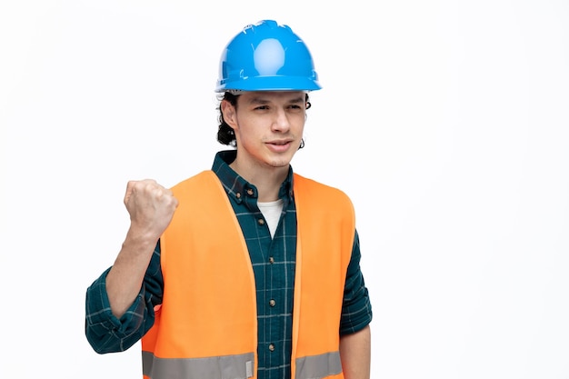 Giovane ingegnere maschio scontento che indossa casco di sicurezza e giubbotto di sicurezza guardando il lato tenendo il pugno in aria isolato su sfondo bianco